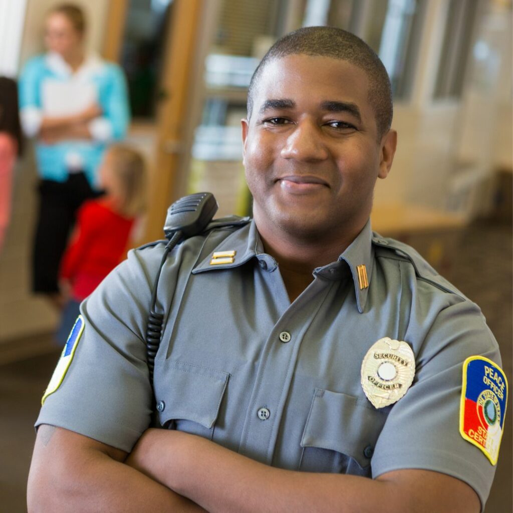 A certified school security officer stands in a hallway with a teacher and students in the background.