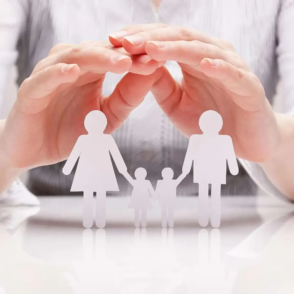 Hands hovering protectively over paper cutouts of a family, including two adults and two children, conveying care and security. The tone is warm and protective.
